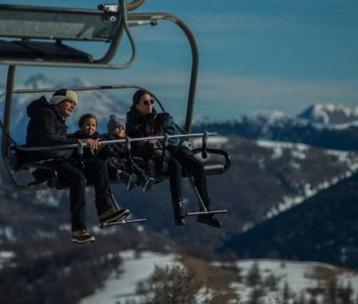 Family having fun on a ski lift with snow-covered mountains in the background during a sunny winter day.